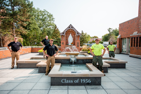 Five men stand by a fountain and statue of Mary. "Class of 1956" is engraved on the fountain's base.