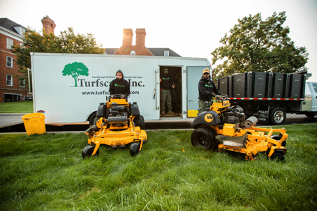 Two Turfscape Inc. employees with yellow mowers stand by their white trailer and a dump truck.