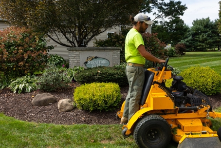 A landscaper on a stand-on mower meticulously mows a lawn next to a garden bed and a sign for "STRATFORD PLACE."