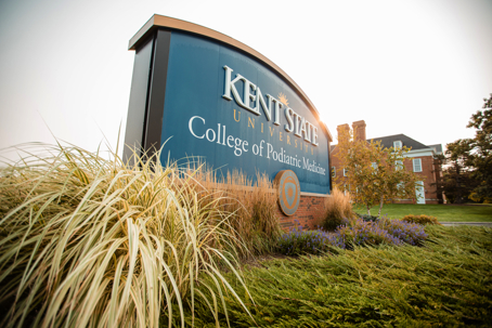 A sunny outdoor view of the Kent State University College of Podiatric Medicine sign, framed by ornamental grasses. Text: KENT STATE UNIVERSITY College of Podiatric Medicine.