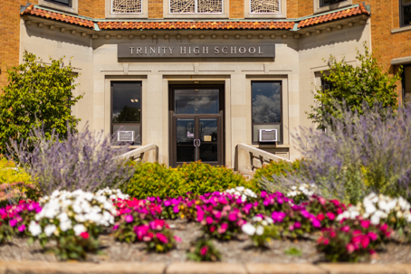 The entrance to Trinity High School is framed by vibrant flower beds with pink, white, and purple blooms in the foreground.
