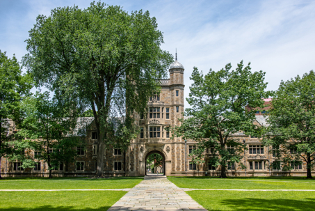 A classic university building with an arched entrance and tower, flanked by mature trees and a green lawn under a bright sky.