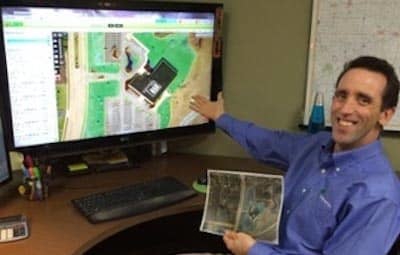 A man in a blue shirt smiles, pointing at a monitor displaying an aerial map. He holds printouts, likely for commercial landscaping bids.