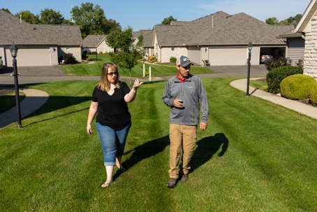 A woman in a black shirt gestures while speaking to a man in a gray uniform and cap, as they walk on a lush green lawn between modern townhomes.