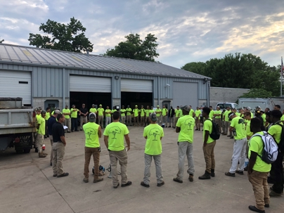 A large group of landscape workers in bright green shirts gather outdoors in front of a service building.