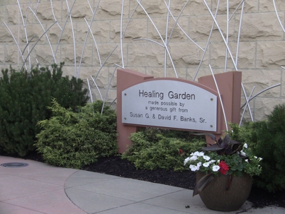 The sign stands amidst green shrubs and a planter with flowers, against a stone wall with metal accents.