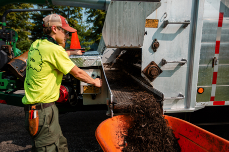 A landscaping worker dispenses dark mulch from a machine into an orange wheelbarrow.