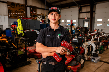 A smiling young technician in a busy workshop holds a power tool, surrounded by various lawn equipment. He is wearing a work shirt and cap.