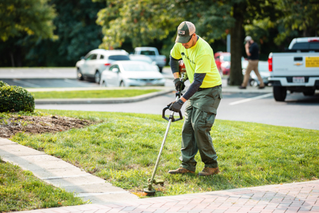 A professional landscaper wearing a bright green shirt uses a string trimmer to edge grass next to a sidewalk.