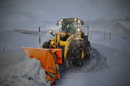 A powerful yellow snow plow with an orange blade pushes a large pile of snow off a wintry road.