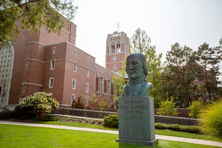 A bronze bust of Archbishop John Carroll (1735-1815) stands on a manicured campus lawn with a large brick building and bell tower in the background.