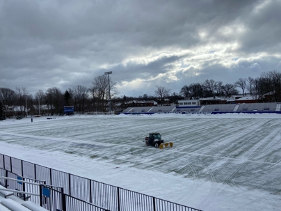 A small utility vehicle with a yellow plow attachment clears light snow from a green turf sports field.