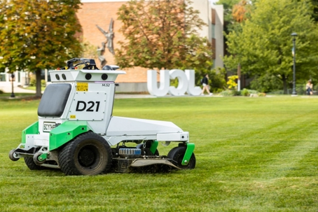 A white and green autonomous Scythe D21 mower cuts a lush lawn on a university campus. In the background, a large UCLA sign