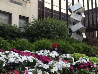 A vibrant university landscape shows colorful flower beds with white, pink, and red blooms in the foreground, with green bushes, a building, and a modern metal sculpture behind.