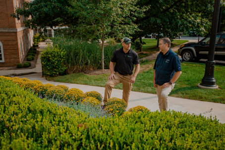 Two men in professional attire stand on a path next to a well-maintained garden with green shrubs and yellow mums.