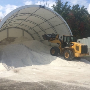 A yellow front-end loader scoops a large pile of de-icing salt from under a white, domed storage facility.