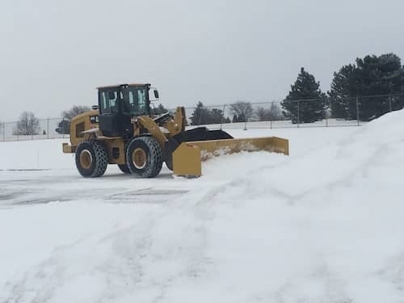 A yellow front-end loader with a large plow attachment clears a snow-covered parking lot.