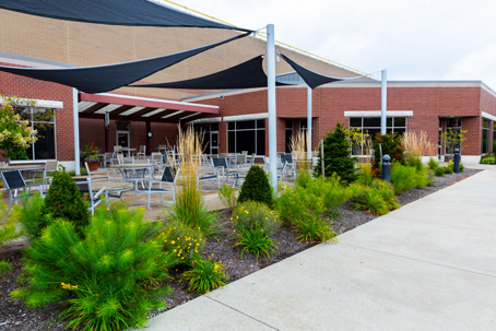 A commercial outdoor patio features a brick building, shaded seating, and vibrant landscaping with diverse plants along a paved walkway.
