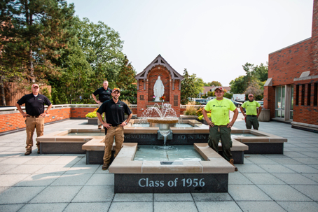 Five landscapers stand proudly around a beautiful multi-tiered fountain featuring a Marian statue. The fountain's base is inscribed with, "Class of 1956".