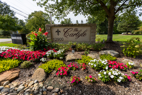 A stone monument for "The Carlyle, 12900" stands amidst a vibrant display of red, white, and green flowers and foliage.