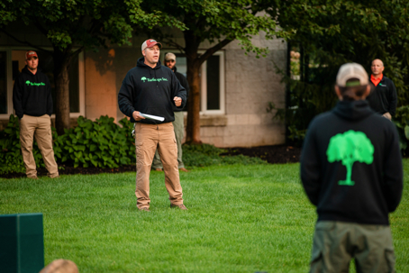 A group of men in black hoodies featuring a green tree logo and khaki pants gather on a lawn.
