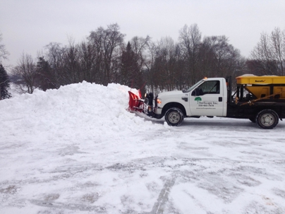 A commercial snow plow truck with a salt spreader pushes a large pile of snow in a parking lot on a winter day.