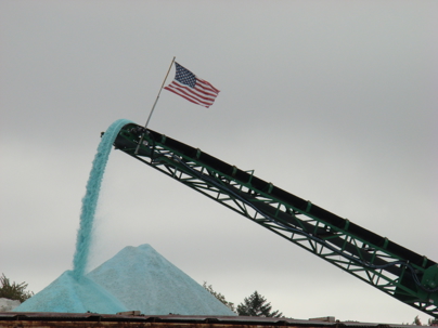 A long conveyor belt deposits light blue road salt into large piles. An American flag flies from the conveyor structure against an overcast sky.