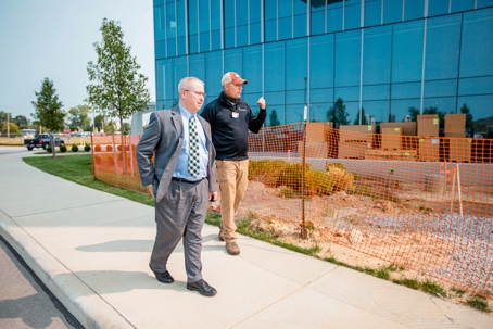 A man in a suit walks alongside a construction worker near a modern glass building and a fenced-off work area.
