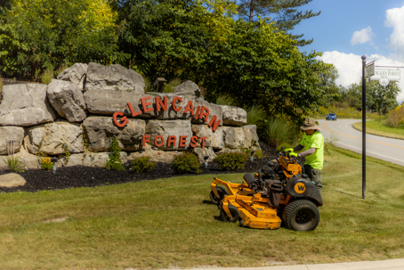 A landscaper on a yellow mower trims grass in front of a stone sign reading, "GLENCAIRN FOREST."